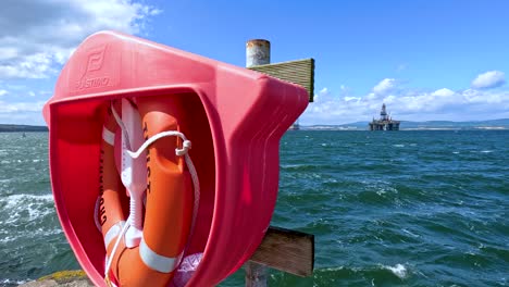 a bright orange lifebuoy ring in a red case is mounted on a wooden pier, with choppy sea and an offshore platform visible under clear daylight. camera pans slightly