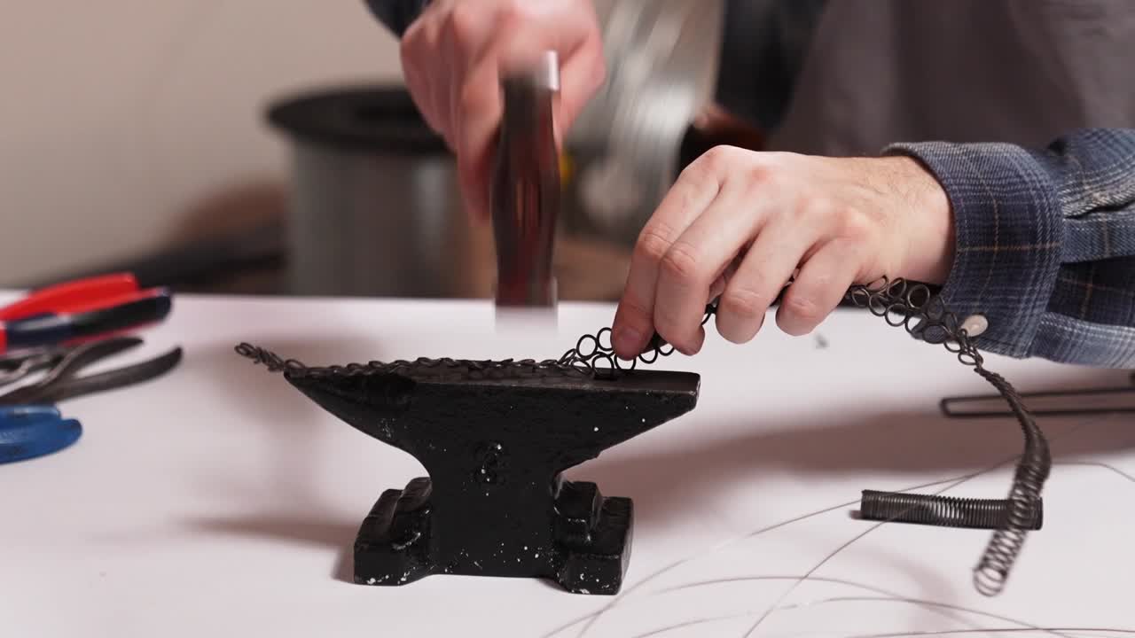 Close up of craftsman shaping metal wire on small anvil using hammer. Traditional Slovak wire craft technique, handmade folk art and heritage craftsmanship in workshop