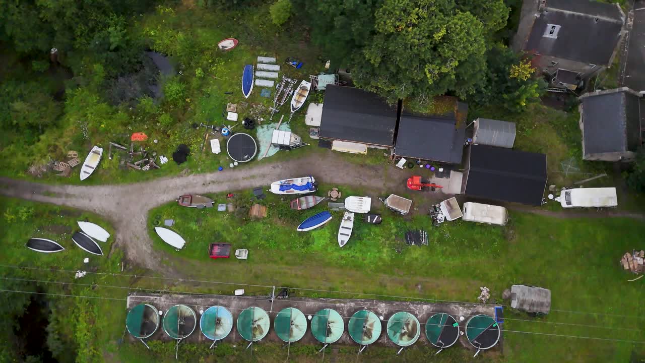 Drone footage glides above a rural boathouse complex with canoes, kayaks, and green tanks, captured in soft daylight with steady overhead movement
