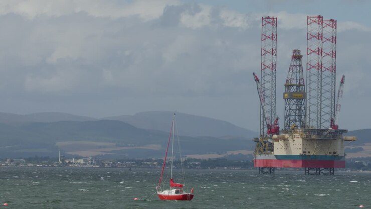 A red fishing boat moves across choppy sea in front of a stationary offshore oil rig, under cloudy daylight with distant hills in Cromarty, Scotland