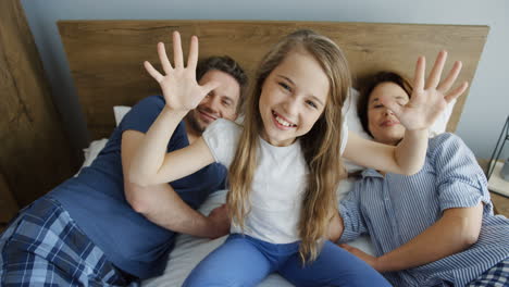 vista superior de una chica rubia sentada en la cama y agitando las manos a la cámara entre sus padres que están mintiendo