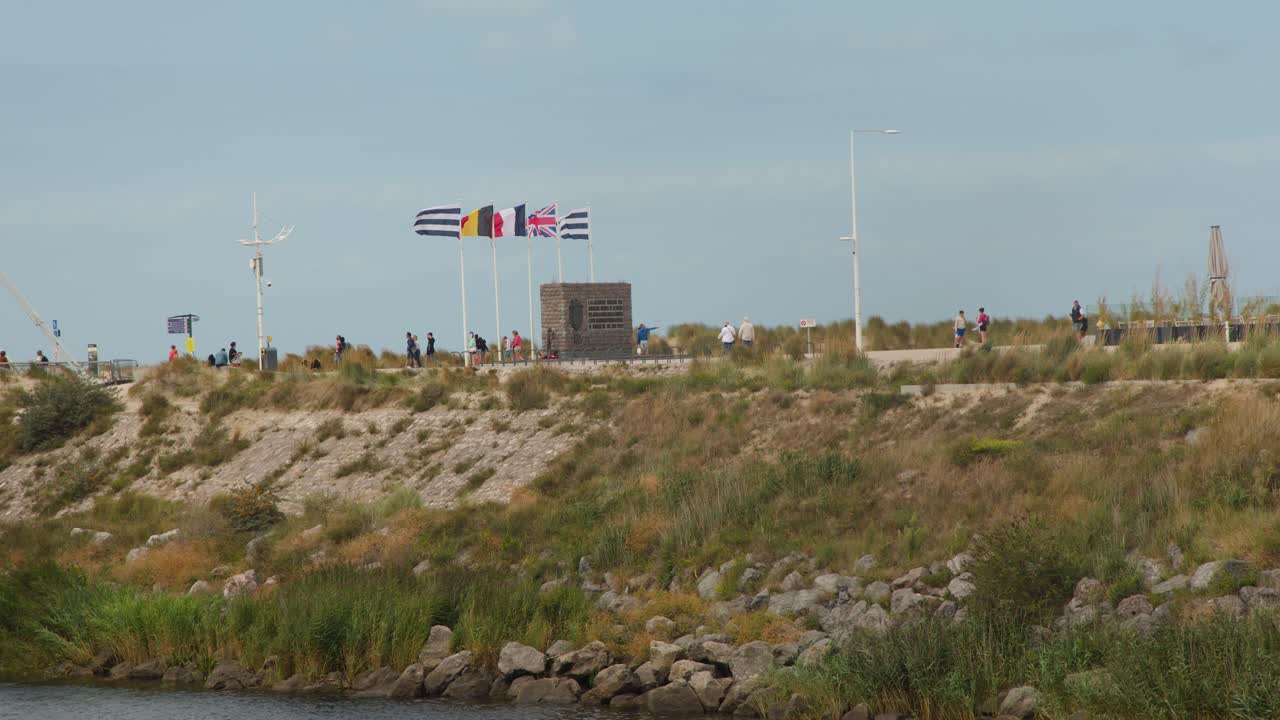 Groups walk past memorial monument and flags on grassy coastal pathway, daylight, wide static shot