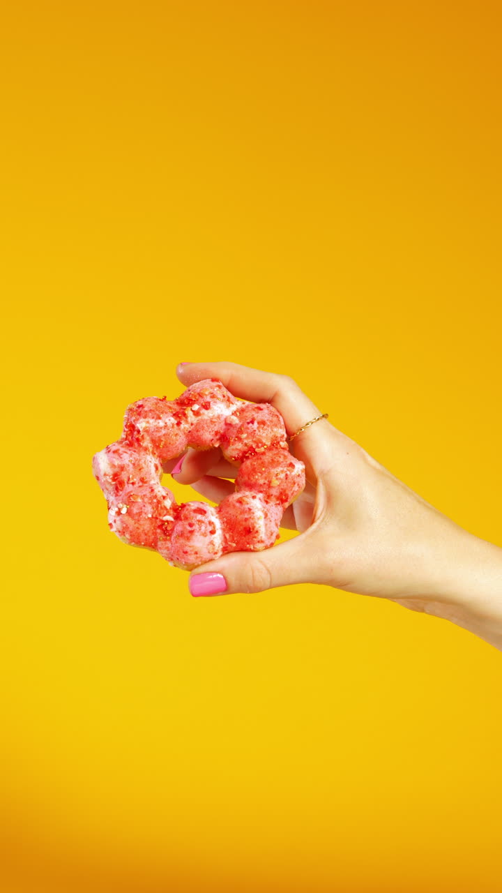 A hand with pink nails enters from the right, holding a bubble-ring donut with white glaze and pink-red topping. The vibrant yellow background adds contrast and energy to the clean minimal composition
