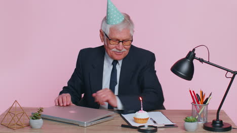 un empresario sénior celebrando un cumpleaños solitario en la oficina, soplando una vela en un pastel pequeño haciendo un deseo.