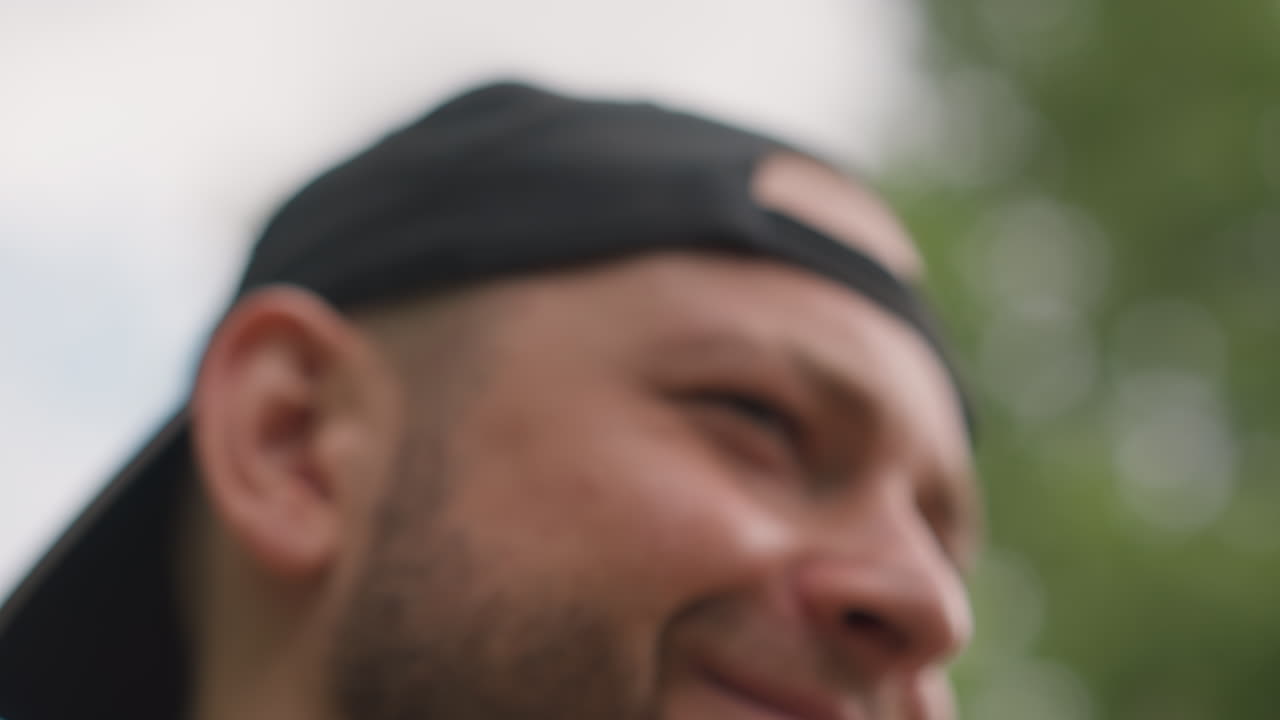 White Man Closeup Smiling With Mischief, Cheeky Performer Reacting To Joke, Tight Portrait, Natural Light Through Trees, Visible Beard And Cap, Emotive Microexpressions, Smug Charm, Authentic Candid