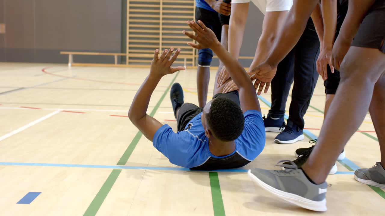 Afroamerikanischer Spieler hält sich vor Schmerz das rechte Knie, während der Trainer die Beweglichkeit auf dem Holzspielfeld testet