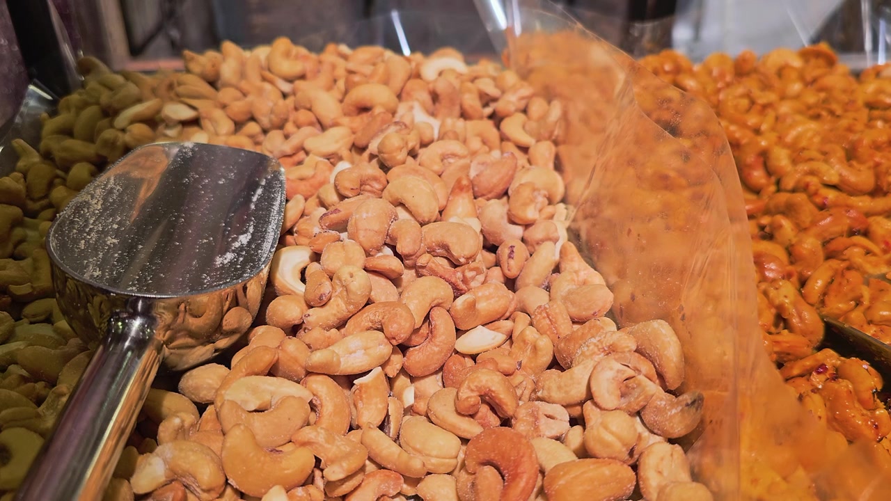Self serve nut bar with cashews, peanuts, almonds, and seasoned mixes arranged in divided bins, with metal scoops, warm shop lighting, and fresh gourmet snack display.