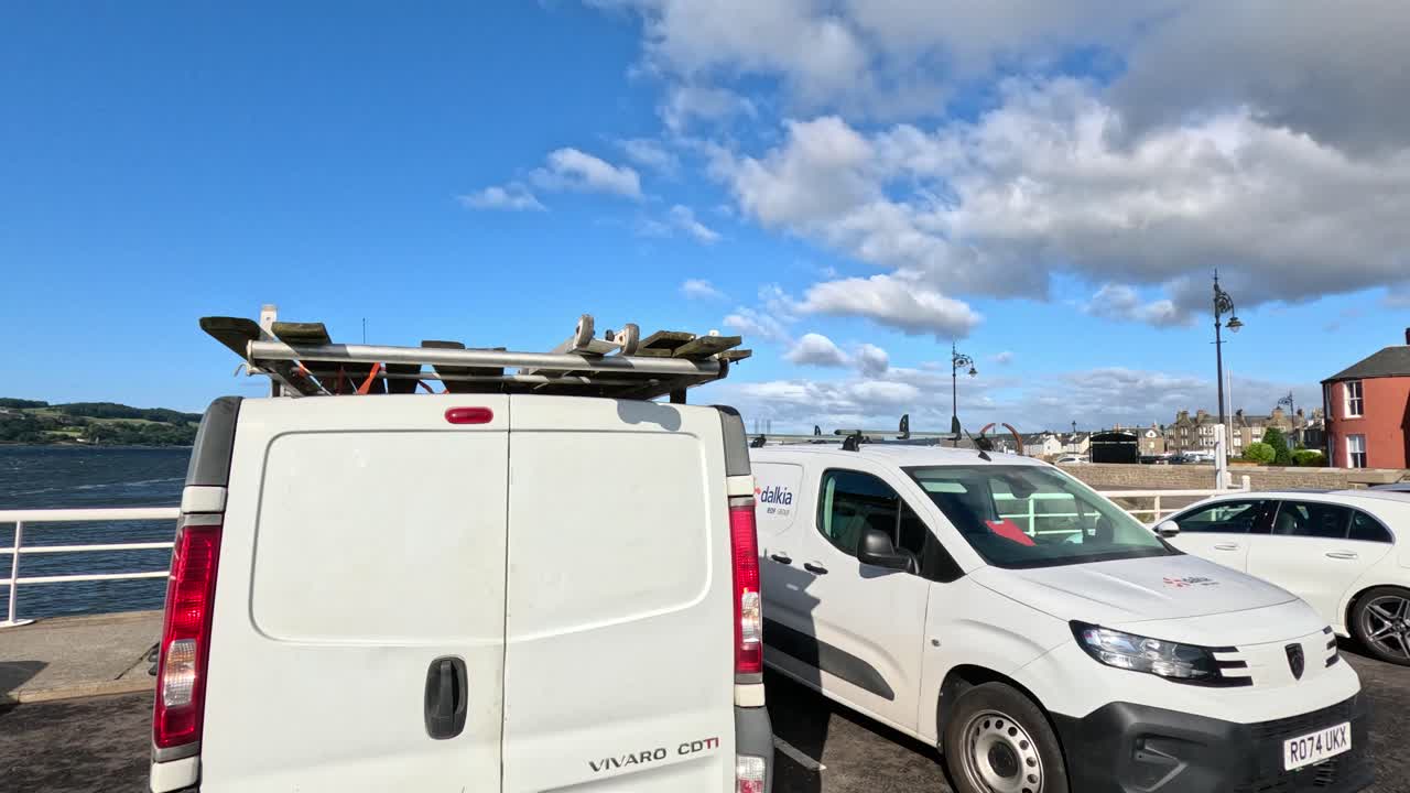 Daytime video pans across a seaside parking area with cars, vans, and a view of the water, under bright blue skies and scattered clouds