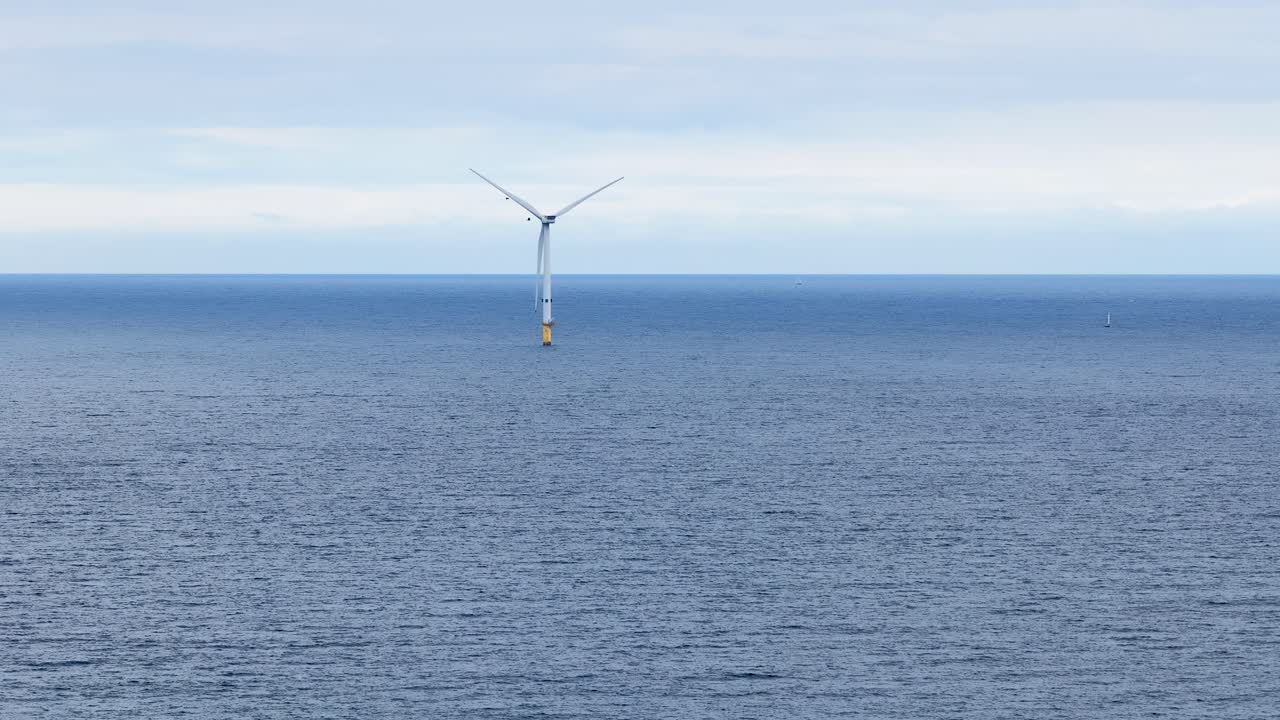 Single offshore wind turbine slowly rotates above calm ocean waters under overcast daylight, captured in a wide, static shot near Whitley Bay, England