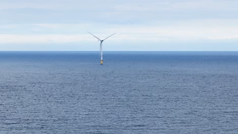 single offshore wind turbine slowly rotates above calm ocean waters under overcast daylight, captured in a wide, static shot near whitley bay, england