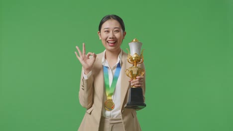 asian business woman in a suit with a gold medal and trophy showing okay gesture and smiling to camera as the first winner on green screen background in the studio