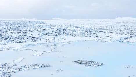 Familia-Caminando-En-La-Orilla-Cubierta-De-Nieve-De-La-Laguna-Azul-En-Islandia,-Antena