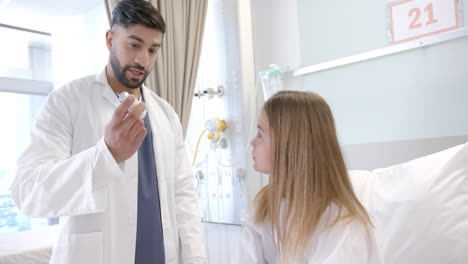 diverse male doctor showing girl patient in hospital bed how to use asthma inhaler, slow motion