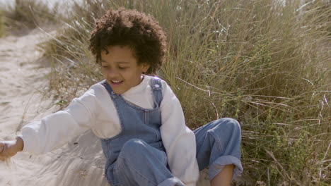 linda niña americana sentada en la playa y jugando con arena en un cálido día ventoso