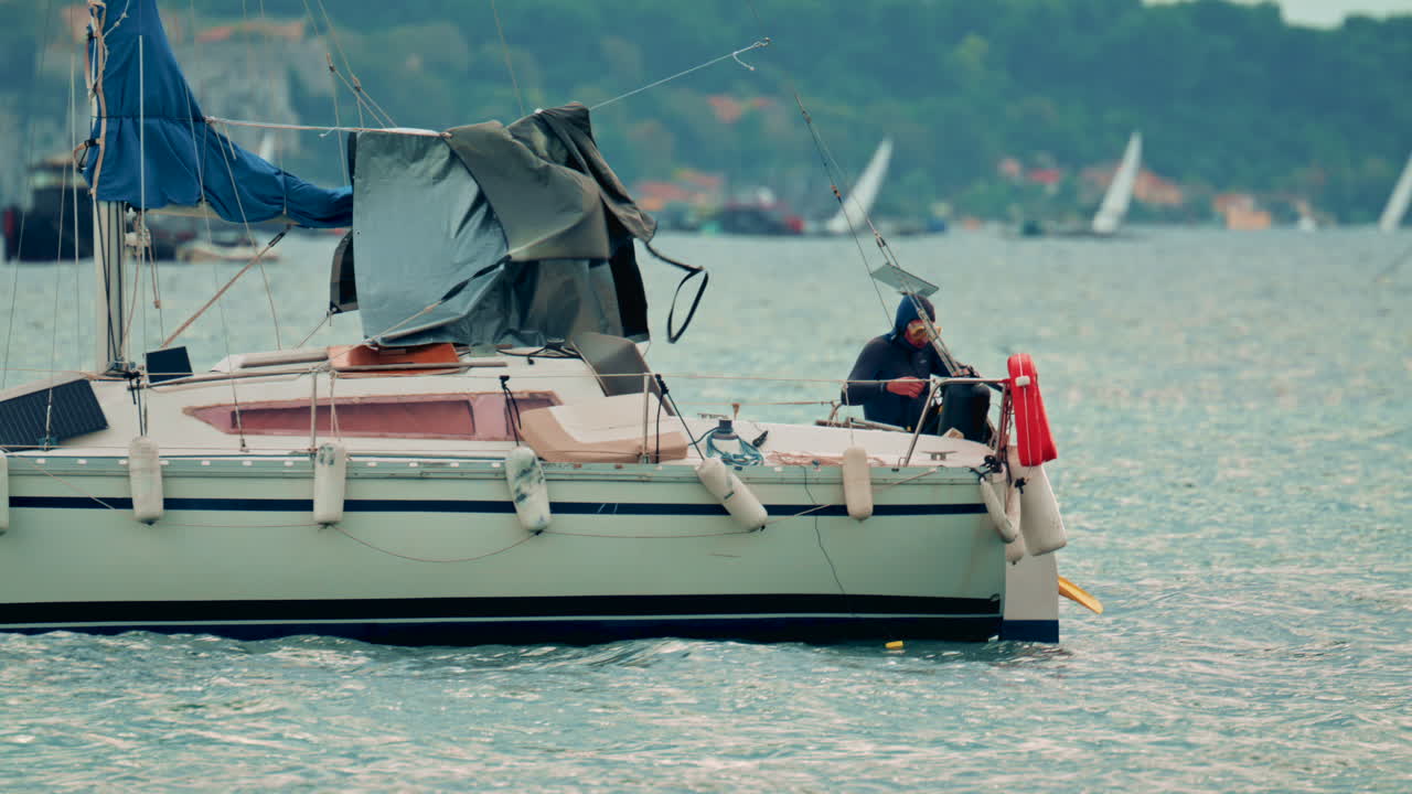 View of a small sailboat rocking on the waves with a lone sailor on deck, diver preparing to jump into sea