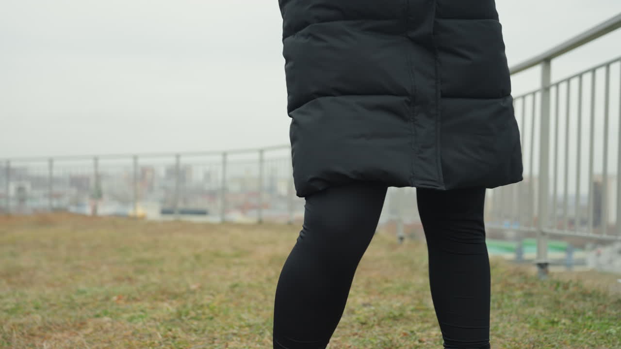 Gros plan sur les jambes d'une femme sud-asiatique dansant sur l'herbe, leggings et bottes noirs, ourlet de doudoune apparent, pas réguliers et rythmés près de la balustrade et du front de mer, texture de l'herbe humide, concentration sur le rythme et l'équilibre.