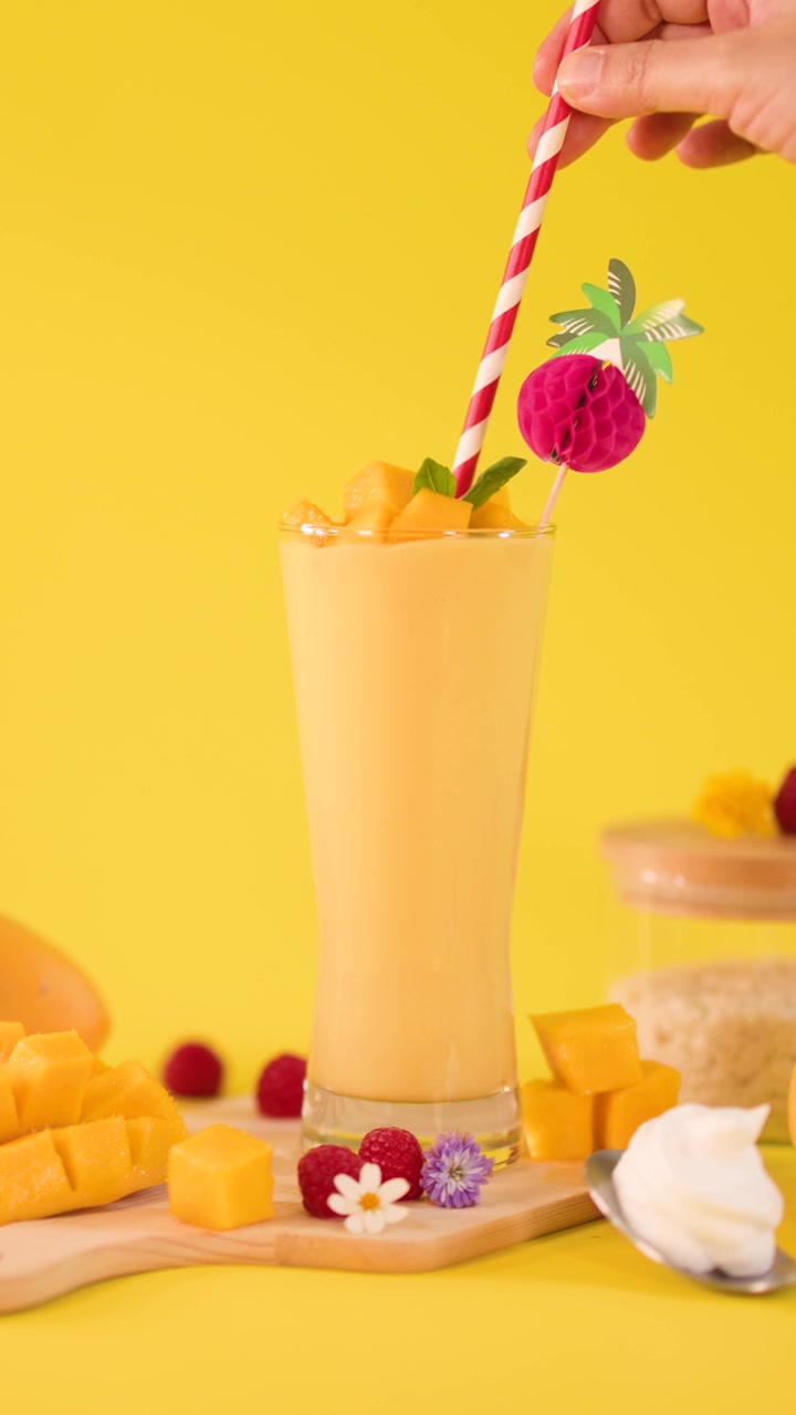 Primo piano di una mano che sistema una cannuccia a righe in un frullato tropicale al mango su uno sfondo da studio giallo brillante