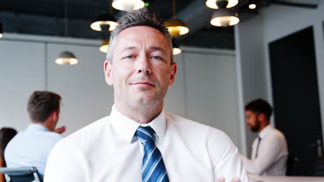 portrait of mature businessman sitting in modern boardroom with colleagues meeting around table in background shot in slow motion