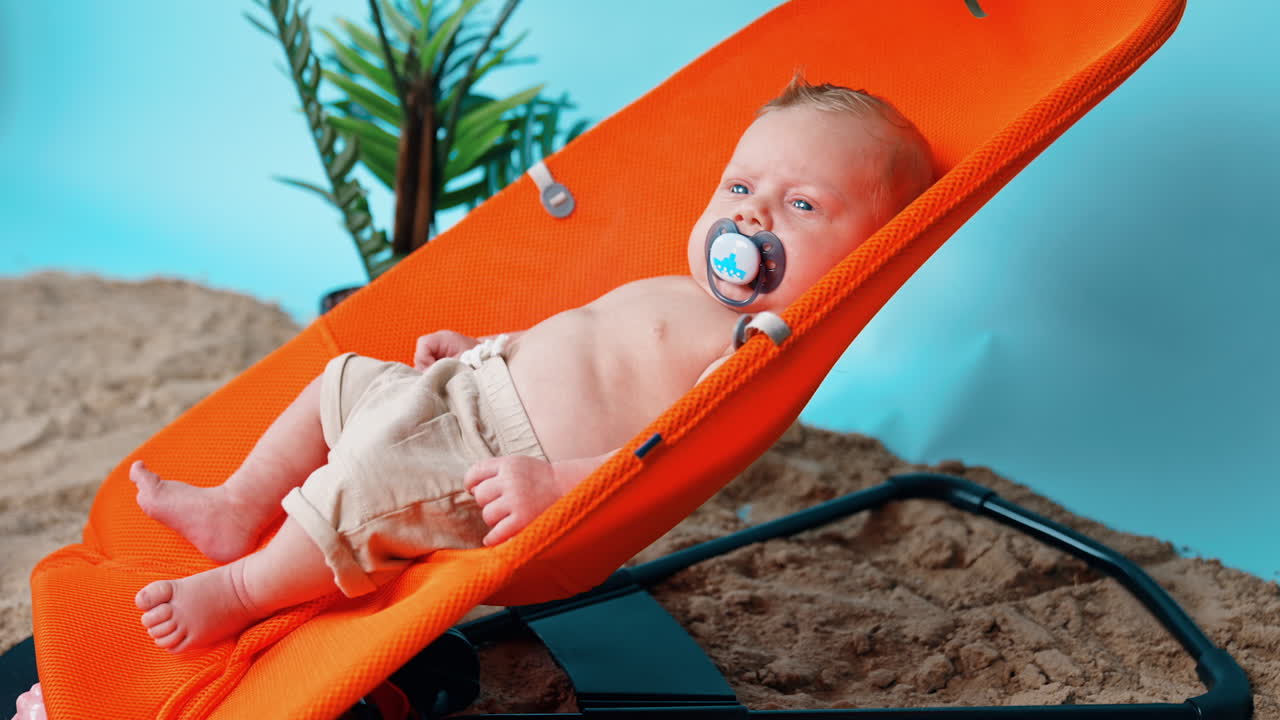 Lovely baby boy with pacifier in mouth lies in chair. Infant is winking from the bright light.