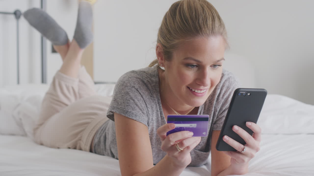 mujer acostada en la cama con teléfono móvil comprando en línea usando tarjeta de crédito con pijama