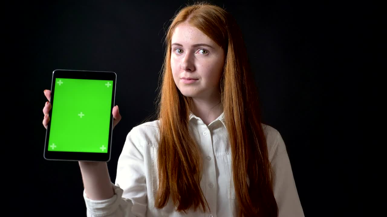 Ginger charming woman typing on tablet and showing display with chromakey into camera, isolated on black background