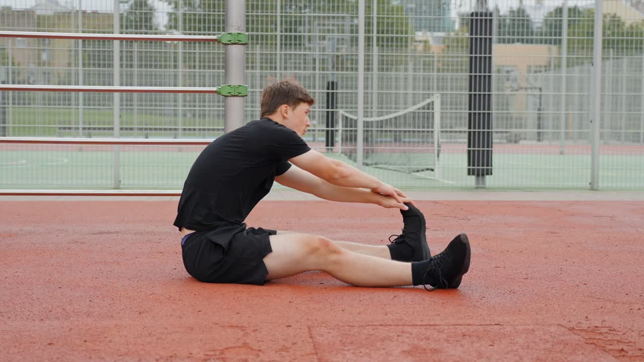 Fixed shot of a young man sitting on the ground in a sports area, stretching forward with straight legs and touching his feet