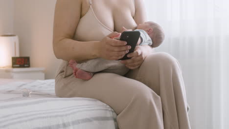 nicht erkennbare mutter stillt ihr neugeborenes baby und benutzt handy, während sie zu hause auf dem bett sitzt