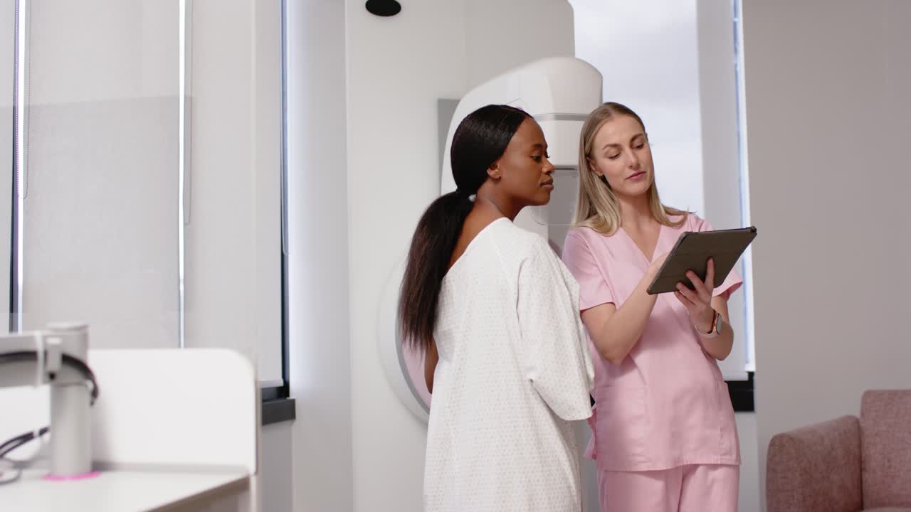 Paziente donna di diverse origini in camice bianco che ascolta, mentre un tecnico tocca il tablet spiegando la mammografia nella sala d'esame.
