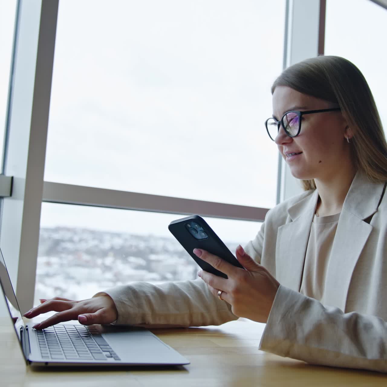 Young woman sitting at desk near the window using gadgets. Working lady uses smartphone for business communication