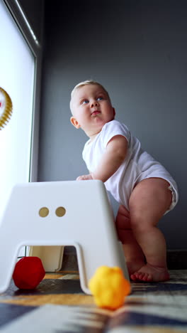 cute caucasian baby boy stands up holding by the chair. one-year-old child learning to stand. low angle view. vertical video.