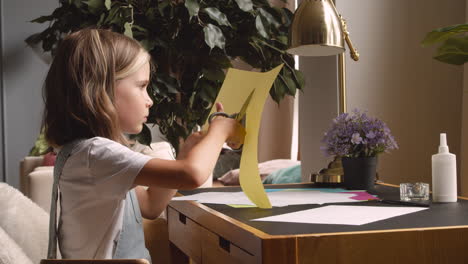 side view of blonde girl cutting cardboard sitting at desk