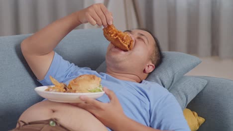 close up of a fat asian man eating fast food having fried chicken while lying on a sofa in the living room at home