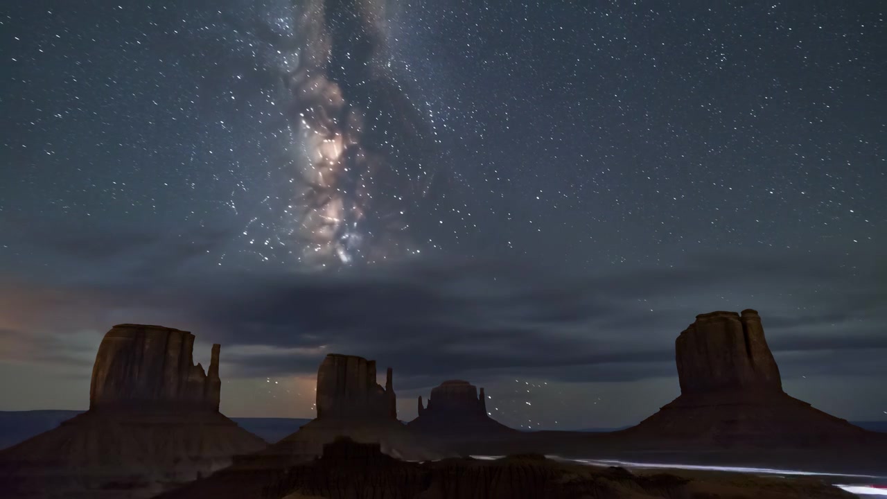 Wide desert scene with isolated sandstone buttes, layered mesas, rugged erosion forms, starry sky, glowing dusk colors, and long cloud band stretching above distant horizon with dramatic depth