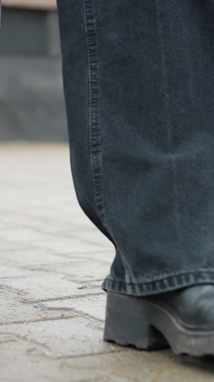 Close up of someone's black trousers and black boots walking slowly toward camera on wet paved urban path, face not visible, capturing calm motion of steps and moody rainy atmosphere