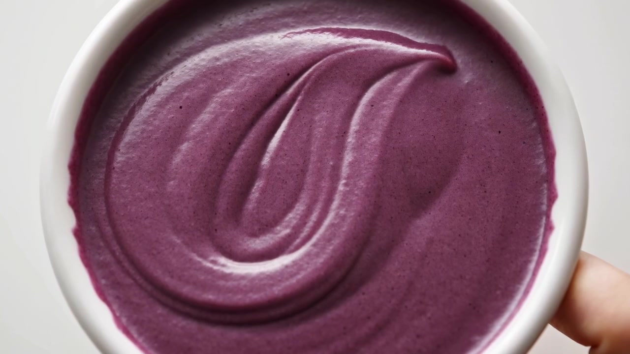 Overhead close-up of spoon lifting and stirring thick purple mixture inside white bowl, leaving smooth curved trails and glossy swirl patterns across creamy surface, with hand steadying bowl and