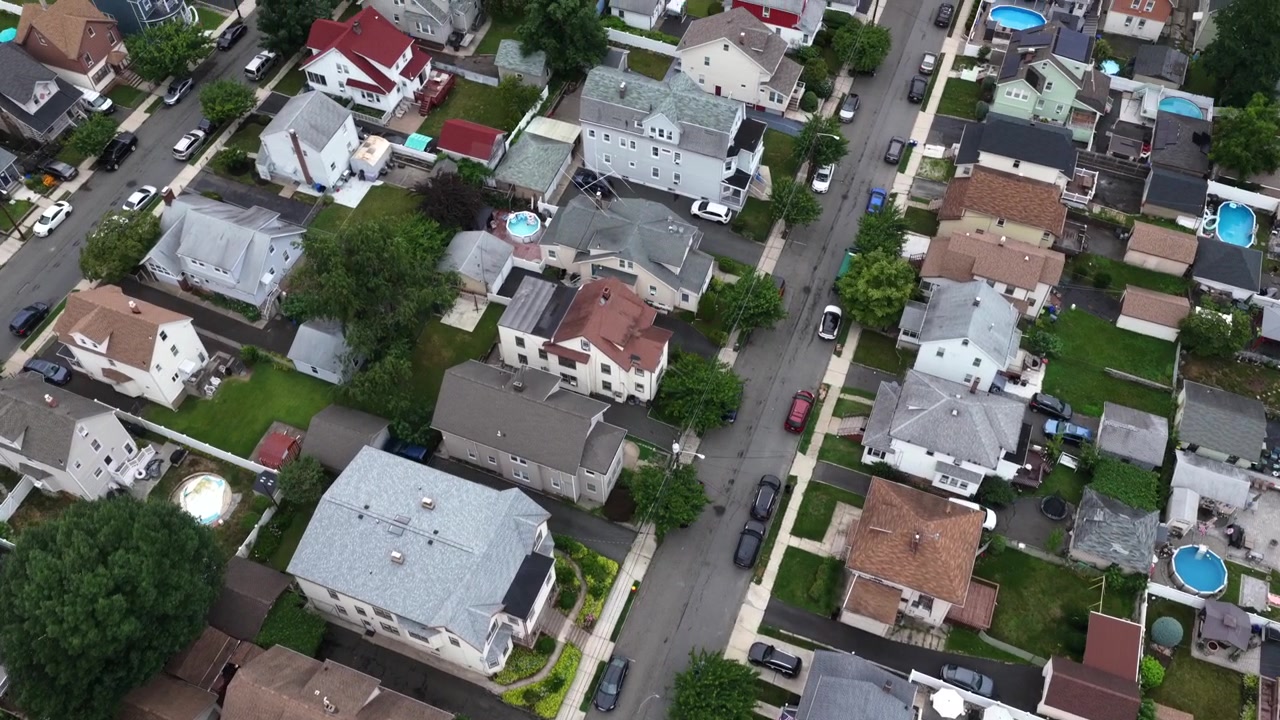 Una strada suburbana si snoda tra una casa rossa e piscine nel cortile a Belleville, New Jersey. Le auto percorrono l'asfalto tra le abitazioni con pannelli solari sui tetti e tetti a capanna. Sorvolo aereo con drone dall'alto