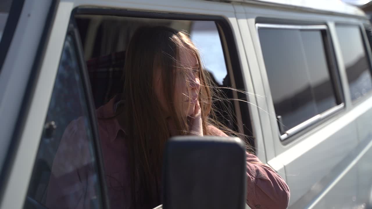 A young long-haired red-haired girl sitting in a caravan by a lake enjoys nature and fresh air.