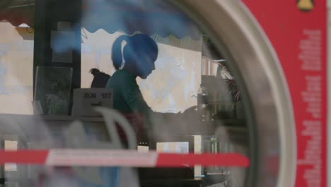 a stationary handheld shot of a reflection from the glass surface of a front-load washing machine shows a sitting woman browsing her phone inside the laundry shop while waiting for her clothes