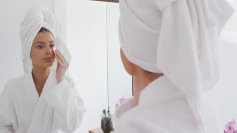 woman in bathrobe removing make up with cotton pad while looking in the mirror