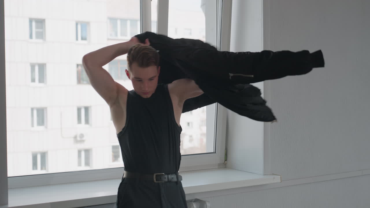 Young man retrieves black jacket from windowsill, slips arms into sleeves and adjusts fit, tall office building visible through window, bright minimalist room with stool and travel bag on floor