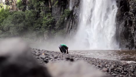 Hombre-Parado-En-El-Río-Debajo-De-Una-Cascada-Lavando-La-Suciedad-Y-La-Mugre-De-Sus-Piernas