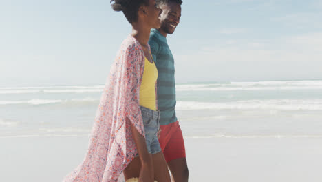 feliz pareja afroamericana caminando y tomándose de las manos en una playa soleada