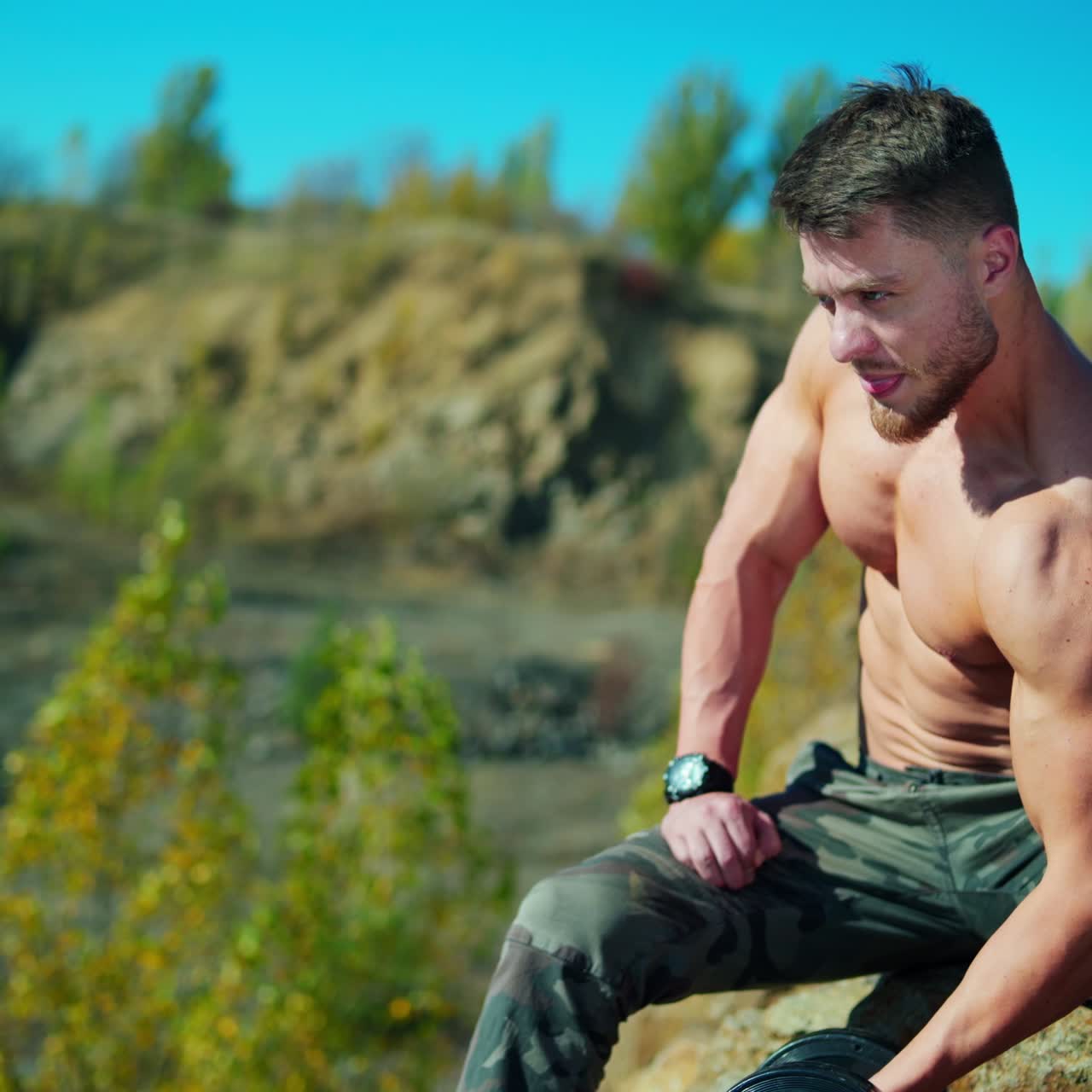 Athletic man pumping arm muscles outdoors. Shirtless bodybuilder sitting on a stone and training with a dumbbell in a sunny day.