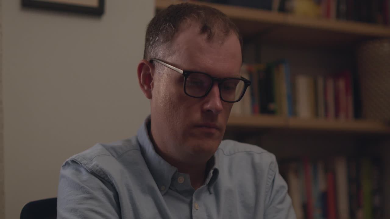 A stressed man in glasses takes off his glasses and rubs his eyes while at work with books in the background