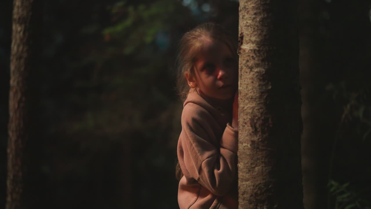 Scared child hides behind tree trunk in night forest. Little girl lost in evening nature park during hiking. Kid threatened by darkness in wild wood