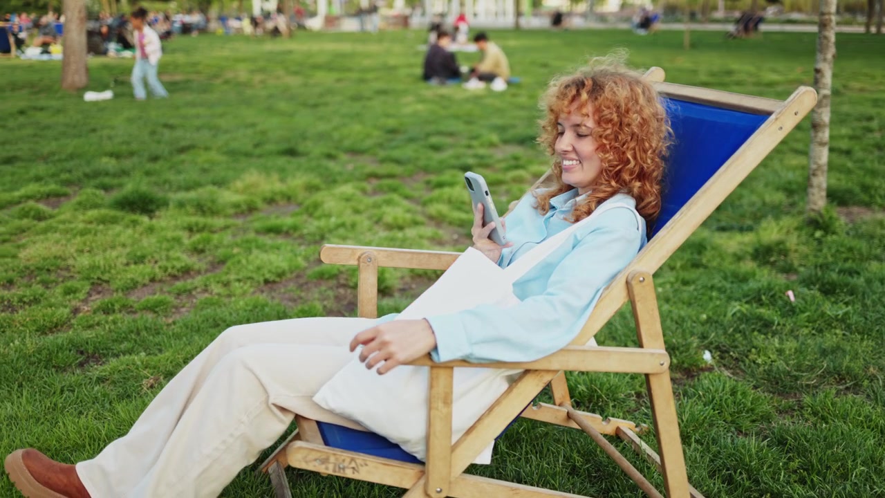 Glad ung kvinde bruger smartphone i en liggestol i parken.