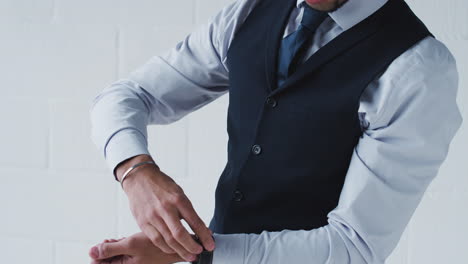 portrait of young businessman in suit fastening wristwatch and tie standing against white studio wall