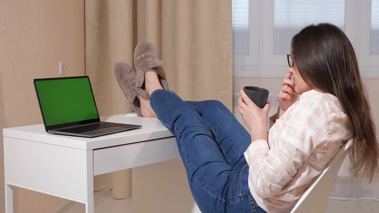 una mujer joven lanza sus piernas sobre la mesa y bebe café mientras mira una computadora portátil con pantalla verde