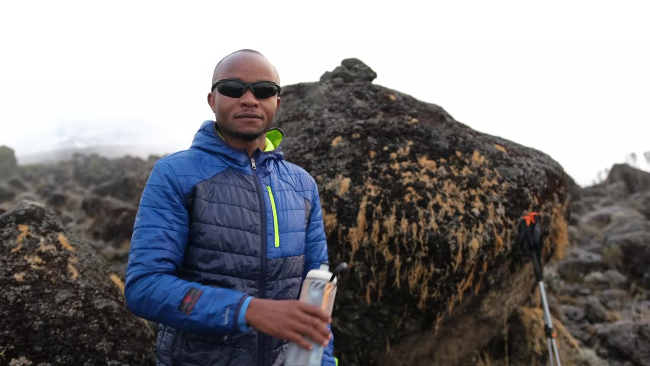 A black man drinks water from a bottle in front of a boulder wearing glasses while trekking