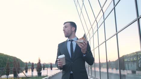 A businessman is eating a sandwich and drinking coffee in front of a building